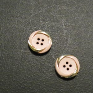 May include: Two decorative buttons with a light pink center and a gold-colored spiral design around the edge. Each button has four small holes in the center for sewing. The buttons are on a dark background.