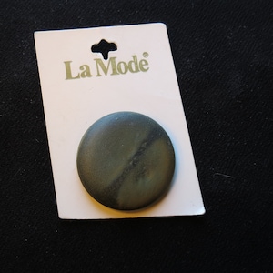 May include: A round, dark green button with a marbled appearance, attached to a white card with the "La Mode" logo. The button is approximately 1.5 inches in diameter. The background is a dark fabric.