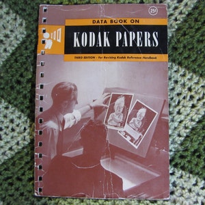 May include: A vintage Kodak Papers Data Book, third edition, with a spiral binding. The cover features the text "KODAK PAPERS" in bold white letters on a black background, with an orange stripe above. The cover also shows a man working with photographs.