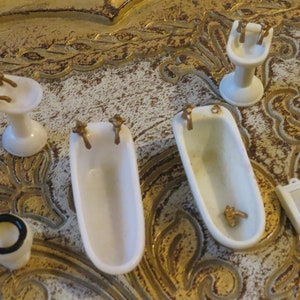 May include: Miniature white bathroom set with gold-colored fixtures. The set includes a bathtub, sink, toilet, and a small mirror. The items are arranged on a decorative surface.