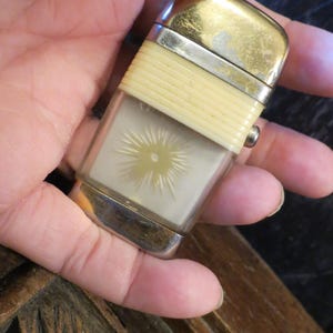 May include: A vintage lighter with a rectangular, clear body and a gold-colored top. The lighter features a cream-colored band and a silver-colored base. The clear body displays a sunburst design.
