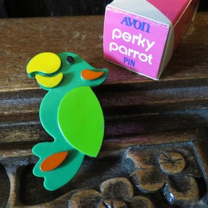 Może przedstawiać: Zielona, żółta i pomarańczowa przypinka w kształcie papugi z napisem "AVON perky parrot PIN" na pudełku. Przypinka w kształcie papugi jest wykonana z tworzywa sztucznego i ma jasny, wesoły design. Przypinka leży na drewnianej powierzchni.