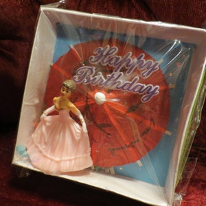 Puede incluir: Una tarjeta de cumpleaños con una figura en miniatura de una mujer con un vestido rosa, sobre un parasol rojo y un fondo azul. Las palabras "Happy Birthday" están escritas en la parte superior.