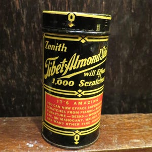 May include: A cylindrical black tin of Zenith Tibet Almond Stick, with gold and red lettering. The label advertises the product's ability to remove scratches from furniture and pianos. The tin is on a wooden surface.