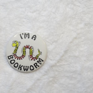 May include: A white button with the text "I'M A BOOKWORM" encircling a cartoon bookworm. The bookworm is yellow, green, and red. The button is on a white, textured surface.