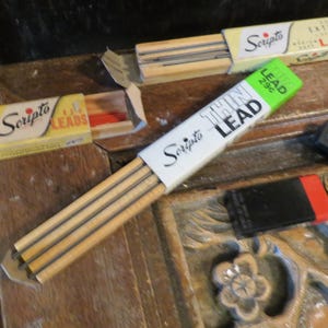 May include: An assortment of vintage Scripto pencil lead refills and erasers. The image shows several boxes of "Extra Thin Leads" and a package of "Thin Lead" refills. Two erasers with red and blue accents are also visible.
