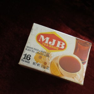 Może przedstawiać: Vintage pudełko MJB Tea Bags z białym i czerwonym logo, z tekstem i ilustracją filiżanki herbaty z herbatą i plasterkiem pomarańczy. Pudełko zawiera 16 torebek herbaty i waży 32.3 g.