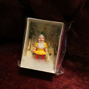 Puede incluir: Figura en miniatura de un gnomo de jardín en una caja. El gnomo viste una túnica amarilla, pantalones rojos y un gorro verde. La caja tiene un fondo de escena de bosque con nieve artificial.