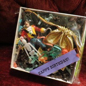 Puede incluir: Una caja de regalo que contiene una figura del Capitán Garfio, una figura de esqueleto y una bolsa dorada con cordón. La caja está llena de papel triturado de colores e incluye una cinta morada con el texto "HAPPY BIRTHAY!".