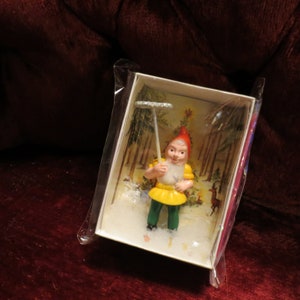 Puede incluir: Figura en miniatura de un gnomo de jardín en una caja. El gnomo lleva un gorro rojo, una camisa amarilla, pantalones verdes y una barba blanca. Sostiene un rastrillo plateado. La caja tiene un fondo de escena de bosque.