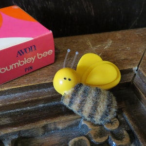 Może przedstawiać: Vintage broszka Avon Bumble Bee z żółtym korpusem i paskowanym, futrzanym brzuchem. Broszka znajduje się obok oryginalnego pudełka, które ma różowy, pomarańczowy i niebieski wzór. Na pudełku widnieje napis "AVON bumbley bee PIN".