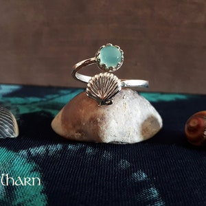 Puede incluir: Un anillo de plata con una piedra preciosa azul claro y un dise&ntilde;o de concha. El anillo est&aacute; sobre una roca.
