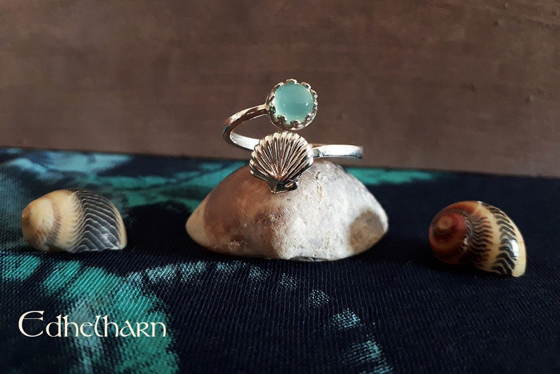 Puede incluir: Un anillo de plata con una piedra preciosa azul claro y un dise&ntilde;o de concha. El anillo est&aacute; sobre una roca.