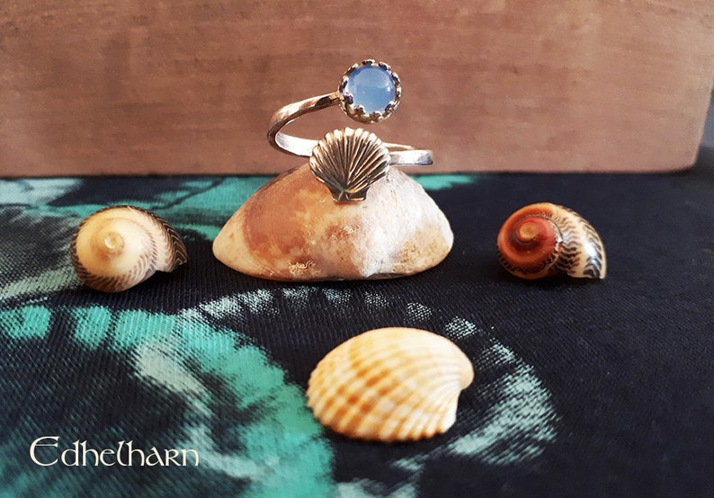 Puede incluir: Un anillo de plata con una piedra preciosa azul y un dise&ntilde;o de concha. El anillo se muestra en una peque&ntilde;a roca con otras conchas.
