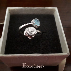 Puede incluir: Un anillo de plata con una piedra preciosa azul y un dise&ntilde;o de concha. El anillo est&aacute; en una caja blanca con la palabra "E&oacute;helbarn" impresa en la parte inferior.