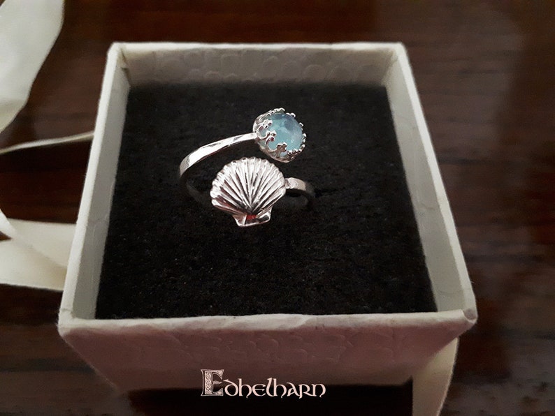 Puede incluir: Un anillo de plata con una piedra preciosa azul y un dise&ntilde;o de concha. El anillo est&aacute; en una caja blanca con la palabra "E&oacute;helbarn" impresa en la parte inferior.
