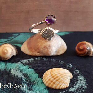 Puede incluir: Un anillo de plata con una piedra preciosa p&uacute;rpura y un dise&ntilde;o de concha. El anillo se muestra en una peque&ntilde;a roca con otras conchas.