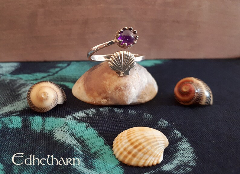 Puede incluir: Un anillo de plata con una piedra preciosa p&uacute;rpura y un dise&ntilde;o de concha. El anillo se muestra en una peque&ntilde;a roca con otras conchas.