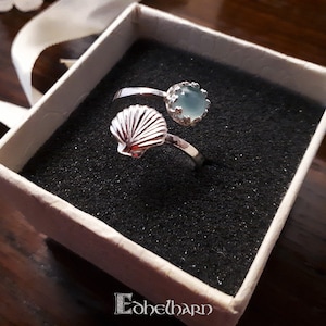 Puede incluir: Un anillo de plata con una piedra preciosa azul y un dise&ntilde;o de concha. El anillo est&aacute; en una caja blanca con forro negro.