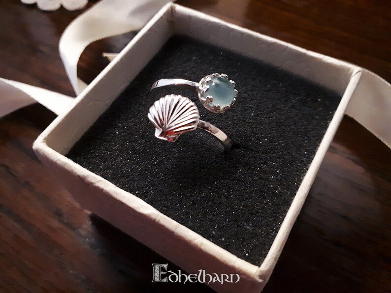 Puede incluir: Un anillo de plata con una piedra preciosa azul y un dise&ntilde;o de concha. El anillo est&aacute; en una caja blanca con forro negro.