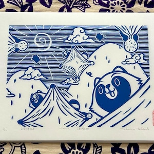 Peut inclure: Gravure sur bois bleue et blanche d'un raton laveur avec une montagne et des nuages en arrière-plan. L'impression est intitulée "Tanuki" et signée "Makana Silva".