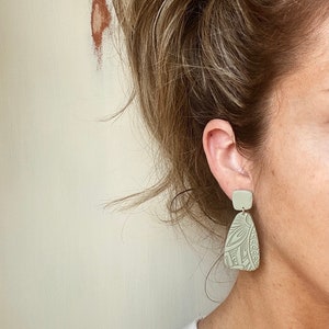 May include: A pair of light green dangle earrings with a geometric pattern. The earrings are made of polymer clay and have a matte finish.