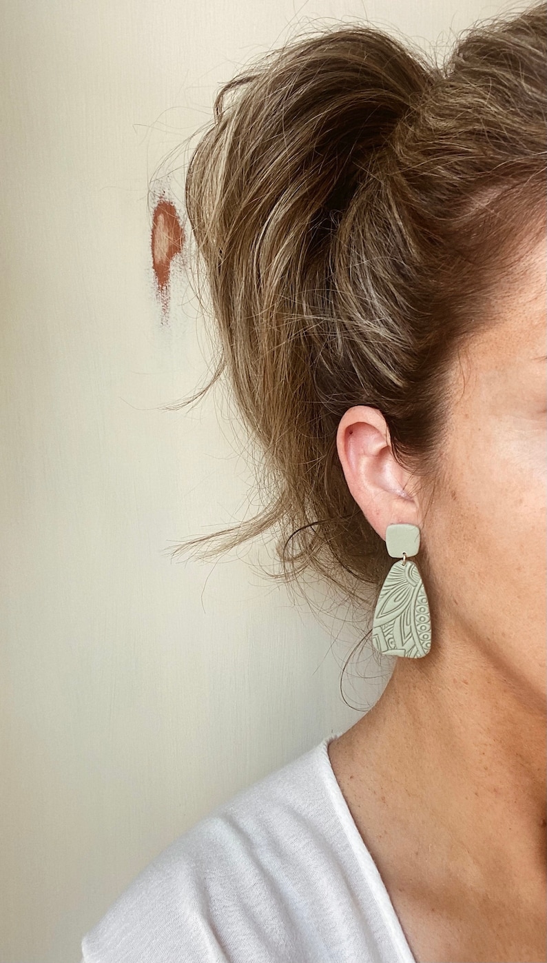May include: A pair of light green dangle earrings with a geometric pattern. The earrings are made of polymer clay and have a matte finish.