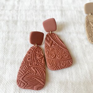 May include: A pair of terracotta-colored dangle earrings with intricate geometric patterns. The earrings are made of polymer clay and have a square-shaped top piece.