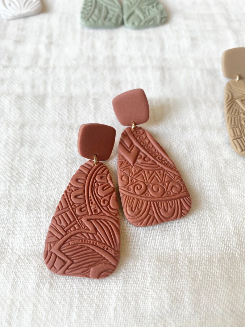 May include: A pair of terracotta-colored dangle earrings with intricate geometric patterns. The earrings are made of polymer clay and have a square-shaped top piece.