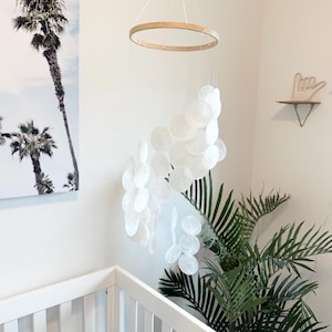 May include: A white baby mobile with circular, shell-like elements suspended from a wooden ring. The mobile hangs above a white cot. A palm tree print and a green plant are in the background.