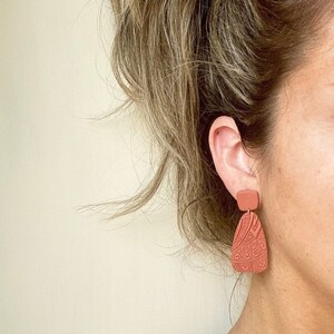 May include: A pair of terracotta-colored dangle earrings with a geometric design. The earrings are made of polymer clay and have a matte finish.
