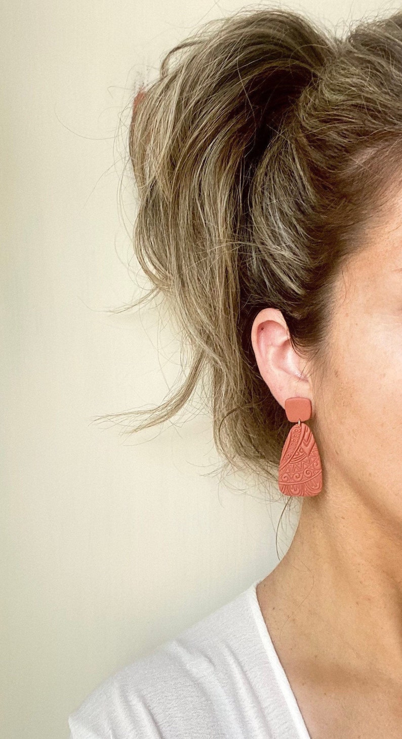 May include: A pair of terracotta-colored dangle earrings with a geometric design. The earrings are made of polymer clay and have a matte finish.