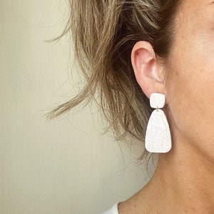 May include: White ceramic earrings with a geometric design. The earrings are dangle style and have a square top piece and a teardrop bottom piece.