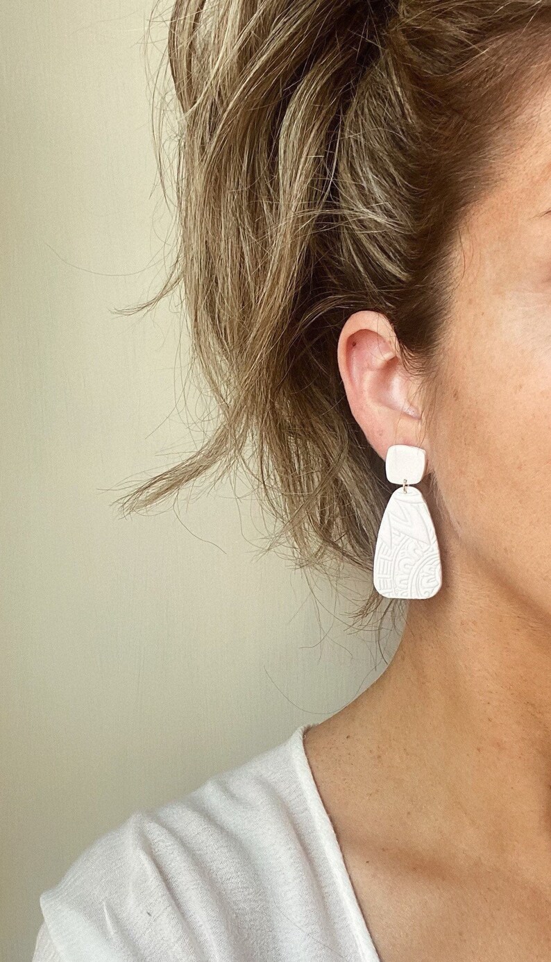 May include: White ceramic earrings with a geometric design. The earrings are dangle style and have a square top piece and a teardrop bottom piece.