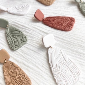 May include: Set of five pairs of dangle earrings with intricate geometric patterns. The earrings are made of clay and come in a variety of colors, including white, brown, green, and red.