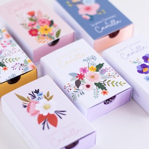 Può includere: Diverse scatole decorative con disegni floreali. Le scatole sono in vari colori, tra cui rosa, blu e bianco, e presentano illustrazioni di fiori e foglie. Il testo "Le premier anniversaire Camilla" è stampato su alcune scatole.
