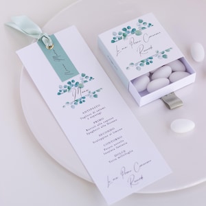 Può includere: Un biglietto di menu bianco e una piccola scatola con il testo "La mia Prima Comunione Ricordo" e un design di foglie di eucalipto. Il biglietto di menu ha un nastro verde acqua e un accento dorato. La scatola contiene caramelle bianche.