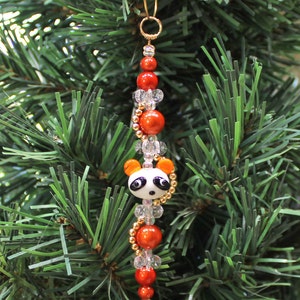 May include: A red and white panda bear Christmas ornament with gold beads and clear glass beads. The ornament is hanging on a green artificial Christmas tree.