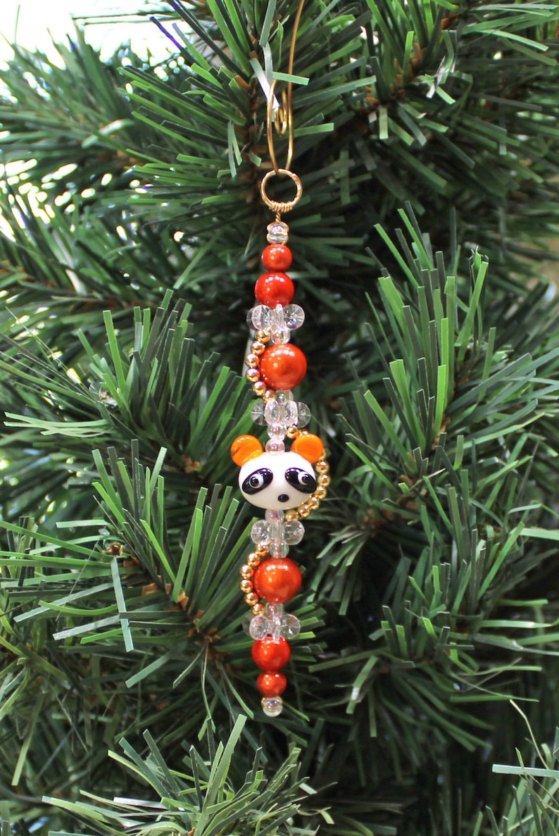May include: A red and white panda bear Christmas ornament with gold beads and clear glass beads. The ornament is hanging on a green artificial Christmas tree.