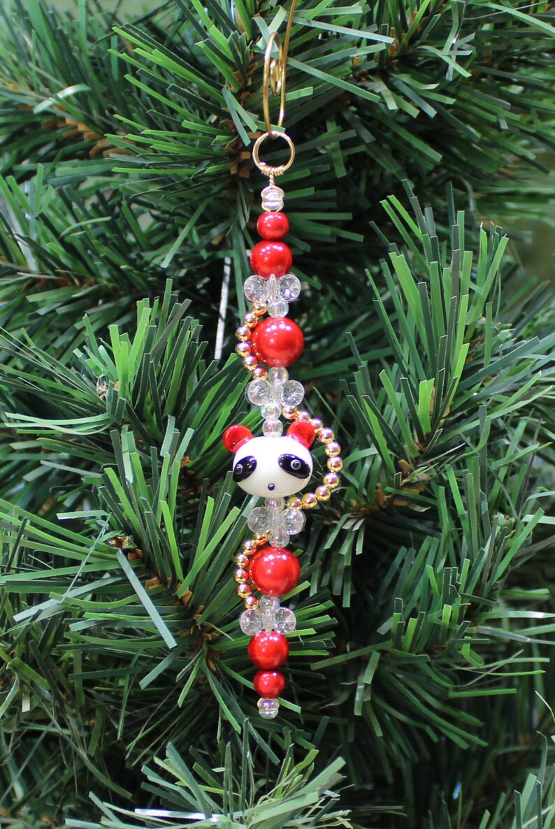 May include: A Christmas ornament featuring a panda bear charm with black and white details, surrounded by red and clear glass beads and gold accents.