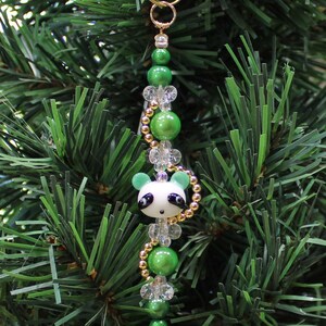 May include: A green and white panda bear Christmas ornament with gold beads and clear beads. The ornament is hanging on a green Christmas tree.