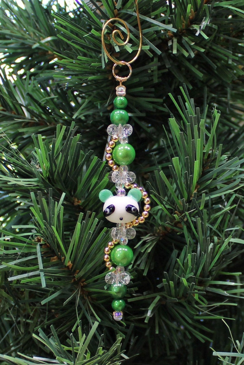 May include: A green and white panda bear Christmas ornament with gold beads and clear beads. The ornament is hanging on a green Christmas tree.