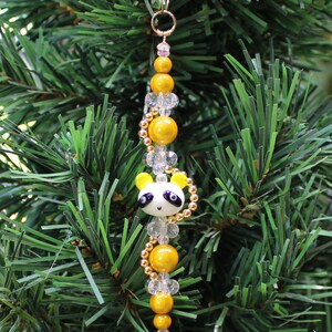May include: A Christmas ornament featuring a panda bear charm with black eyes and a white face. The charm is strung on a gold wire with yellow and clear beads.