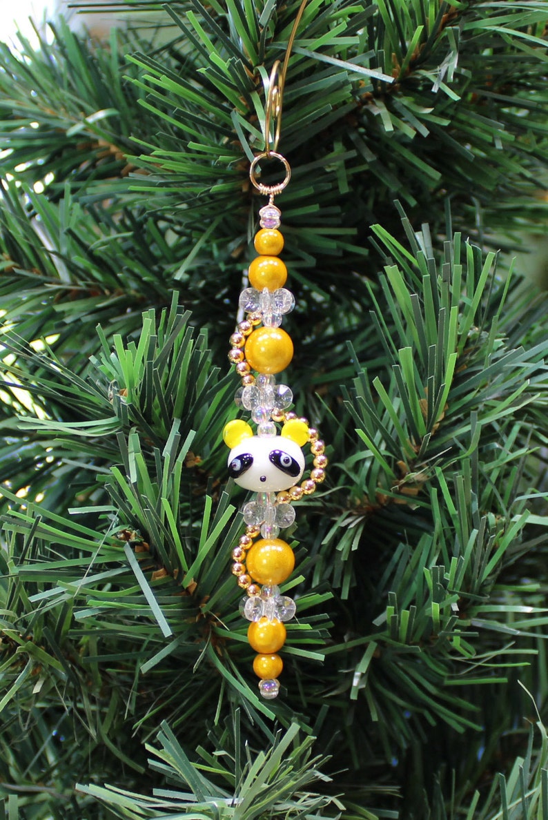 May include: A Christmas ornament featuring a panda bear charm with black eyes and a white face. The charm is strung on a gold wire with yellow and clear beads.