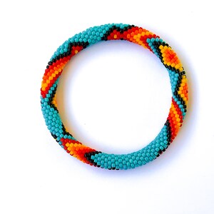 May include: A turquoise, orange, yellow, and black beaded bracelet. The bracelet is a tube shape, with a repeating pattern of colours. The bracelet is approximately 6.35 cm in diameter.