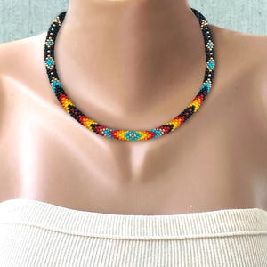 May include: A beaded necklace featuring a repeating geometric pattern of small black, turquoise, yellow, orange, and red glass beads. The circular design is constructed from tiny, uniform seed beads arranged in a continuous, flexible rope-like strand.