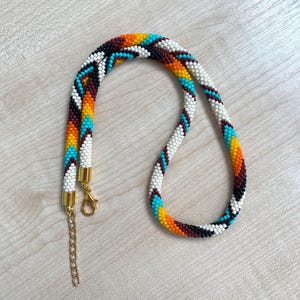 May include: A tubular beaded necklace featuring a repeating chevron pattern in white, black, orange, and turquoise glass beads. The necklace includes gold-toned metal end caps, a lobster claw clasp, and a short adjustable chain extender at the end.