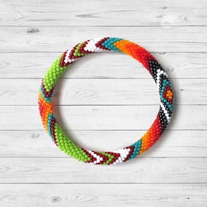 May include: A colorful beaded bracelet in a circular shape. The bracelet features a pattern of green, red, white, orange, black, and turquoise beads. The bracelet is set against a white wooden background.
