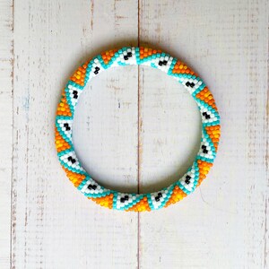 May include: A circular bangle bracelet crafted from small glass beads in a repeating geometric pattern. The design features alternating segments of bright orange, turquoise, and white beads, with small black accents embedded within the white sections.