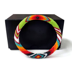 May include: A circular bangle bracelet crafted from small glass beads in a vibrant pattern of lime green, bright orange, red, white, turquoise, and black. The beadwork features geometric shapes and repeating color blocks throughout the band.